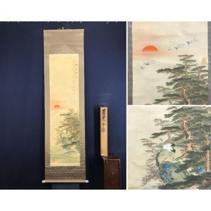 May include: A traditional Japanese scroll painting depicting a landscape with a red sun, pine trees, and flying cranes. The artwork features a serene scene with soft colors and detailed brushstrokes. The scroll is displayed with a wooden box.