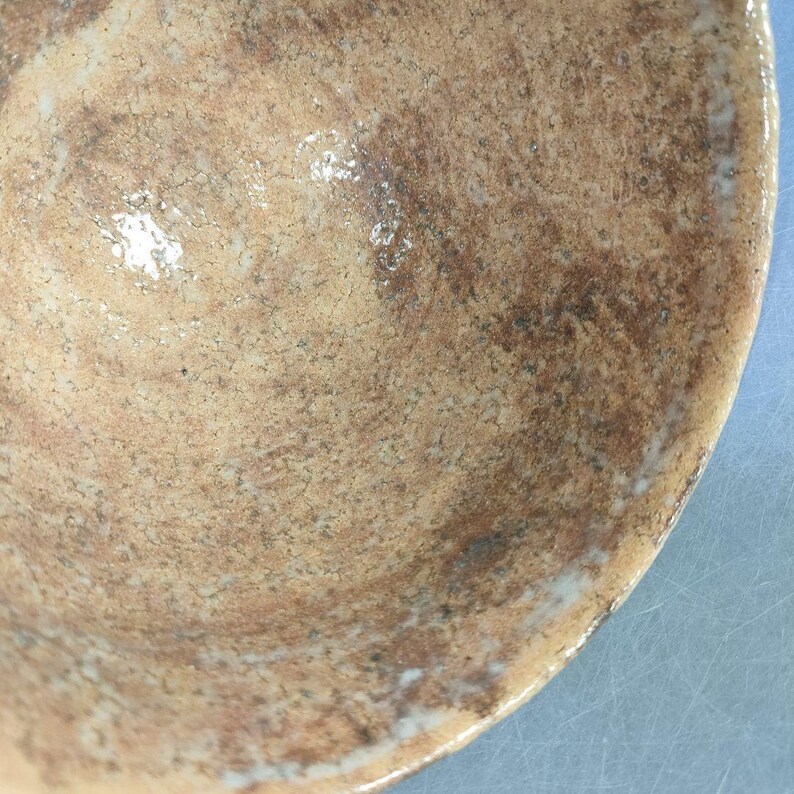 May include: Close-up of a handmade ceramic bowl with a textured surface. The bowl features a blend of brown and tan hues with darker brown marbling. The rim has a lighter, off-white color.