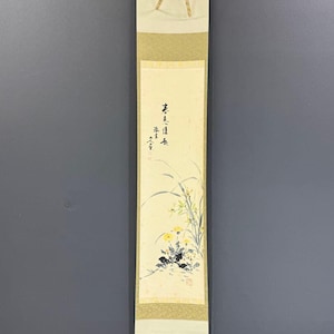 May include: A vertical hanging scroll featuring a painting of yellow flowers and green foliage on a cream-colored background. The artwork includes black ink calligraphy characters and a red seal stamp. The scroll has gold-toned fabric borders.