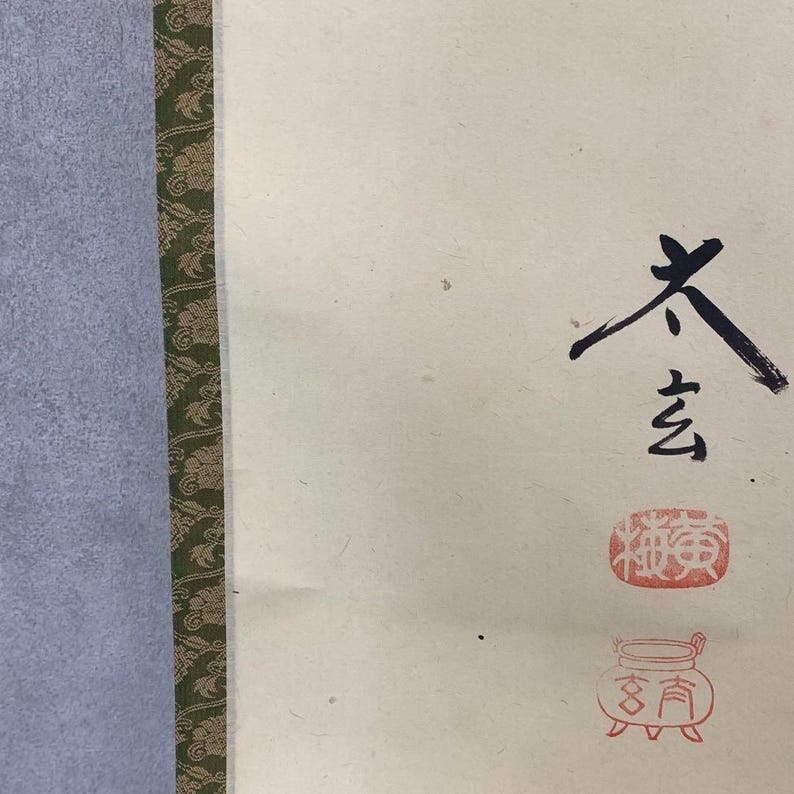 May include: Close-up of a traditional Japanese artwork. The artwork features black brushstrokes of Japanese characters and red stamps on a cream-colored background. A green and gold patterned border is on the left side.