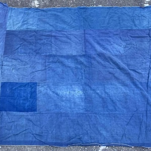 May include: A blue textile with a patchwork design. The fabric is a deep indigo colour, with lighter blue patches sewn together. The edges are frayed, giving it a vintage or handmade appearance. The textile is laid out on a rough, grey surface.