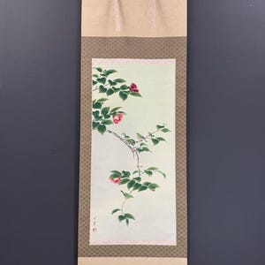 May include: A vertical hanging scroll featuring a painting of camellia branches with green leaves, pink flowers, and two small birds. The artwork is set against a pale green background, framed by brown patterned fabric borders and light beige ends.