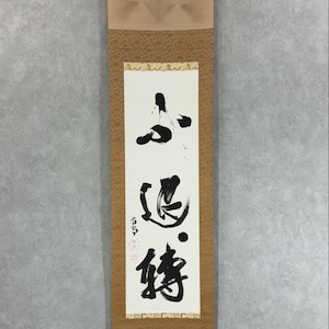 May include: A vertical scroll featuring Japanese calligraphy in black ink on a white background. The scroll is encased in tan paper with decorative accents and a hanging loop. The calligraphy includes abstract figures and characters.