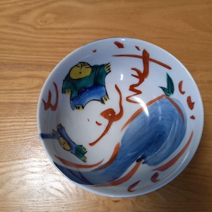 May include: A white ceramic bowl with hand-painted illustrations. The bowl features figures in blue and green, with orange and red brushstrokes. A large blue shape and calligraphy-style text are also present. The bowl is on a wooden surface.