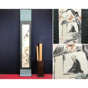 Op de afbeelding: Een Aziatisch schilderij op rol met een landschapsscène en een figuur in een hut. Het schilderij heeft een blauw-witte rand en wordt gepresenteerd met een houten kist en twee houten latten. Het kunstwerk is in verticaal formaat.