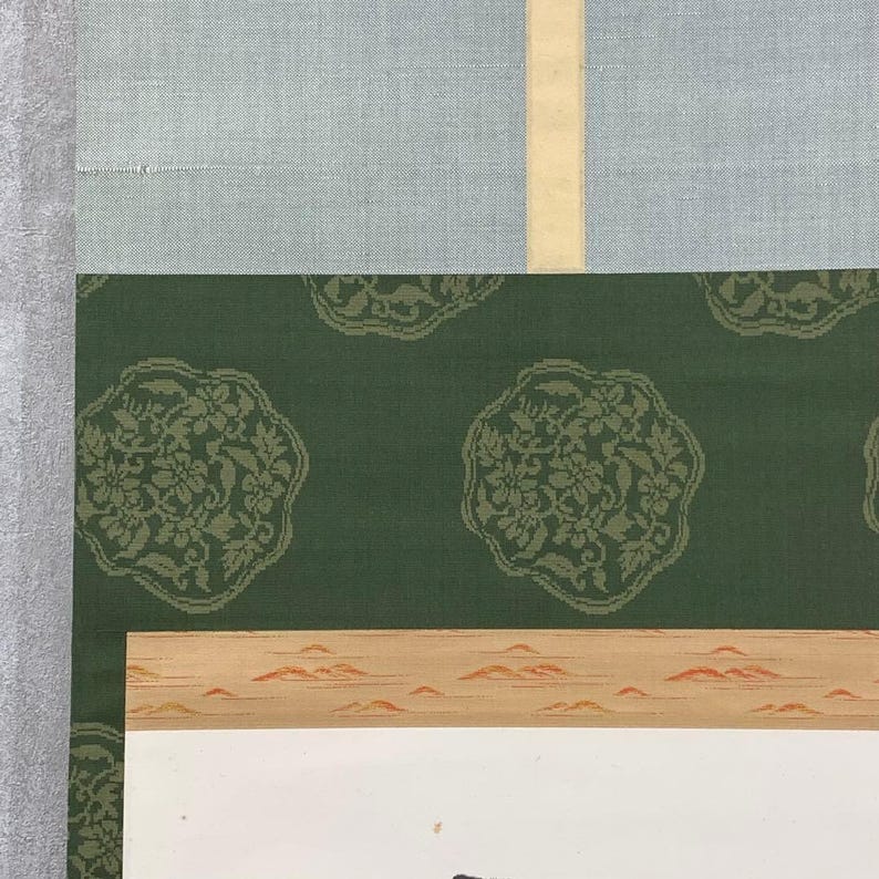 May include: A section of a traditional Japanese scroll painting. The top portion features a light blue fabric, followed by a green fabric with a floral pattern. The bottom section has a landscape painting with a white background.