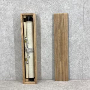 May include: A Japanese scroll in a wooden box. The scroll is rolled up and secured with a black and white patterned band and a decorative cord. The box is made of light-colored wood, and the lid is separate.