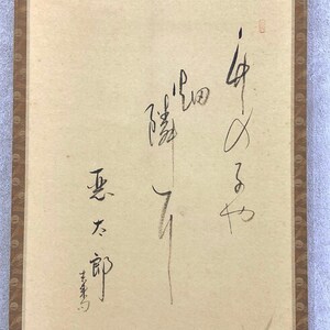 May include: A vintage Japanese scroll with calligraphy in black ink on aged paper. The scroll features vertical text and a brown border. The paper has a light beige hue.