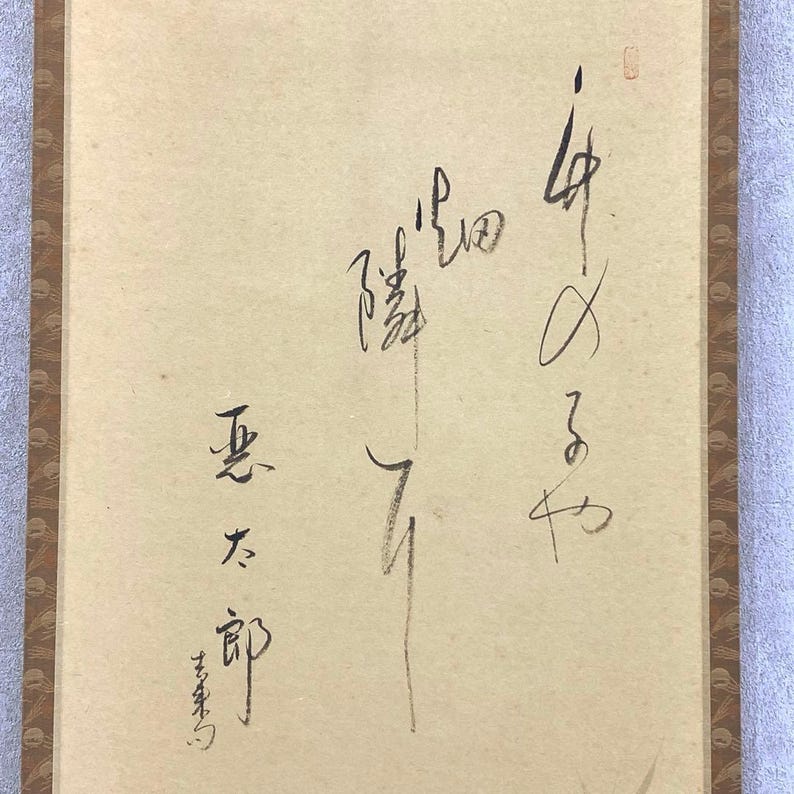May include: A vintage Japanese scroll with calligraphy in black ink on aged paper. The scroll features vertical text and a brown border. The paper has a light beige hue.