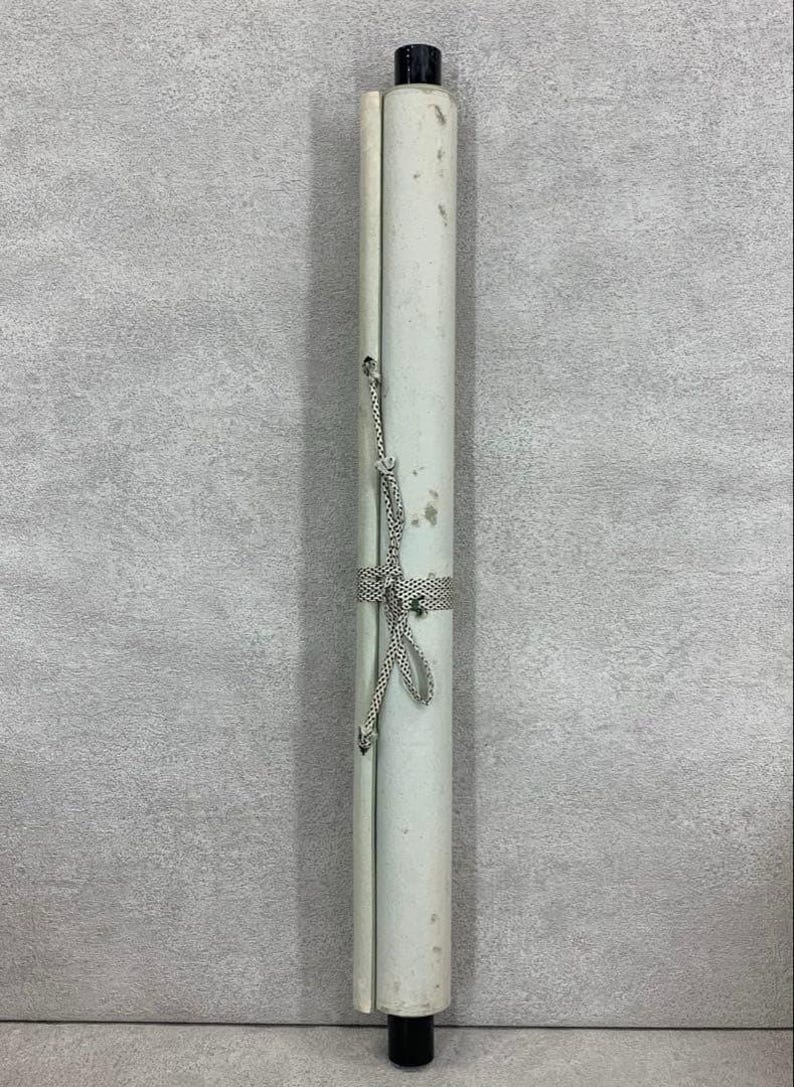 May include: A rolled-up scroll with a white paper surface, secured with a patterned ribbon and cord. The scroll has black caps on both ends. The paper shows signs of age, with some discoloration.