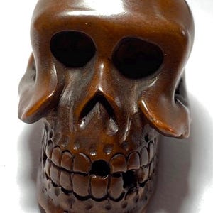 May include: A detailed, carved wooden skull with a polished, brown finish. The skull features hollow eye sockets, a prominent nose cavity, and a detailed row of teeth. The craftsmanship highlights the texture and form of the skull.