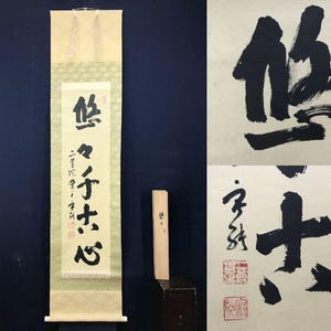 May include: A vertical scroll with Japanese calligraphy in black ink, featuring a central text panel and decorative top and bottom sections. A wooden tool is positioned to the right. Two additional calligraphy pieces are also shown.