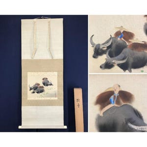 May include: An Asian-style scroll painting featuring a watercolor scene of water buffalo with riders. The artwork is in shades of gray, brown, and blue. The scroll is cream-colored with decorative hanging cords and a wooden storage box.