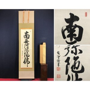 Op de afbeelding: Een verticale rol met Japanse kalligrafie in zwarte inkt, met de zin "Namu Amida Butsu". De rol heeft een beige achtergrond met een goudkleurig koord. Een houten doos en een apart kalligrafiekunstwerk zijn ook zichtbaar.