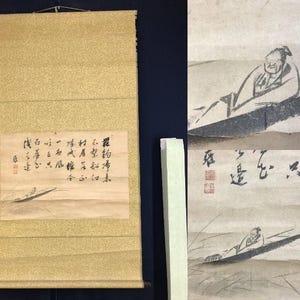 May include: An Asian scroll painting with a gold fabric backing. The artwork features a small boat with a person and calligraphy. The painting is in shades of black, white, and tan.