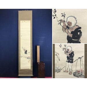 May include: A vintage Japanese scroll painting depicting a figure in a white hat and black clothing, carrying a basket. The artwork uses muted tones, with a focus on the figure and floral elements. The scroll is displayed against a blue wall.