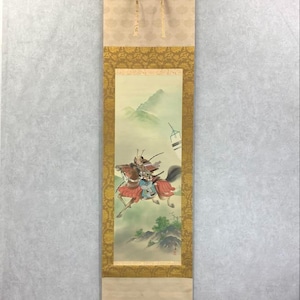 May include: Japanese scroll painting showing a samurai warrior on horseback. The artwork features a detailed illustration of the warrior in armour, riding a brown horse, set against a backdrop of mountains and a pagoda. The scroll has a gold border and a cream-coloured top.