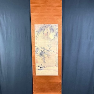 以下が含まれることがあります： 風景画が描かれた伝統的なアジアの巻物。満月、枝、テキストが特徴です。巻物は温かみのあるオレンジ色で、金属製のハンガーから吊り下げられています。