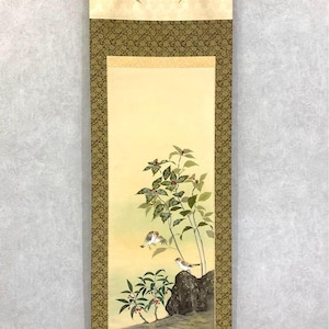 May include: A traditional Japanese scroll painting depicting birds on a rock near a plant with red berries. The artwork has a light yellow background and is framed with a decorative border. The scroll is hung with gold ribbons.