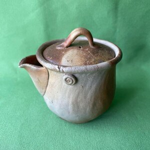 Könnte beinhalten: Eine handgefertigte Keramik-Teekanne mit Deckel und Ausguss. Die Teekanne ist eine Mischung aus Beige und Braun mit einem Spiralmuster an der Seite. Der Griff auf dem Deckel ist gebogen. Die Teekanne ist zum Brühen von Tee bestimmt.