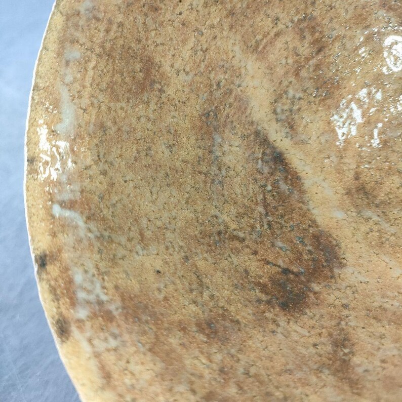 May include: Close-up of a ceramic object with a textured, mottled surface. The colors range from light tan to dark brown, with areas of white and darker spots. The object's edge is visible, suggesting a rounded shape.