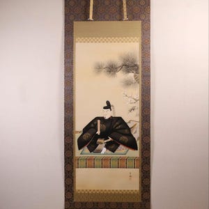 May include: A Japanese scroll painting featuring a seated figure in black robes, holding a scroll. The artwork is framed by a patterned border in shades of brown and gold. The background depicts a tree and a light-colored sky.