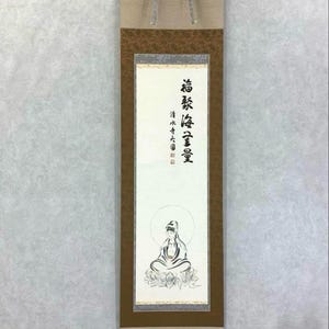May include: A traditional Japanese scroll painting featuring a seated figure with a halo, likely a Buddhist deity. The artwork is in black and white with Japanese calligraphy. The scroll has a neutral color palette with tan and brown accents.