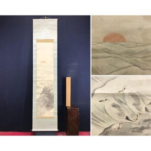 May include: A vertical Japanese hanging scroll featuring an ink wash painting of two seagulls perched on a rocky shore with a setting sun over rolling waves in the background. The scroll is presented with a light blue border and a wooden storage box.