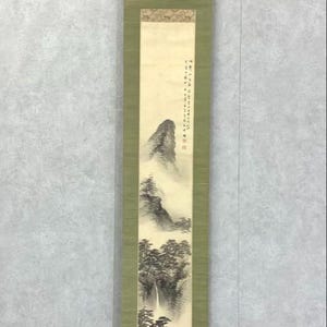 May include: A vertical, traditional Asian scroll painting. The artwork depicts a mountain landscape in shades of black, gray, and white. The scroll is framed by a light green fabric and has wooden dowels at the top and bottom. The painting features a waterfall and trees.