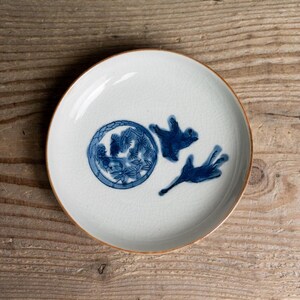 May include: A white ceramic plate with a blue and white floral design. The plate has a brown rim and is set against a wooden background. The plate's design includes a circular motif and two abstract shapes.