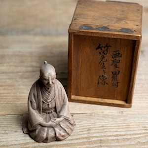 Könnte beinhalten: Eine sitzende, grau getönte Figur einer älteren Person in traditionellen Gewändern, die Hände im Schoß gefaltet. Daneben eine Holzkiste mit japanischen Schriftzeichen. Die Figur und die Kiste stehen auf einer Holzoberfläche.