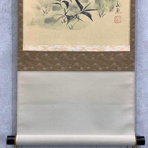 May include: Japanese scroll painting featuring a bamboo and foliage scene in muted green and gray tones. The artwork is mounted on a cream-colored paper with decorative borders in brown and gold. The scroll is finished with black wooden dowels.