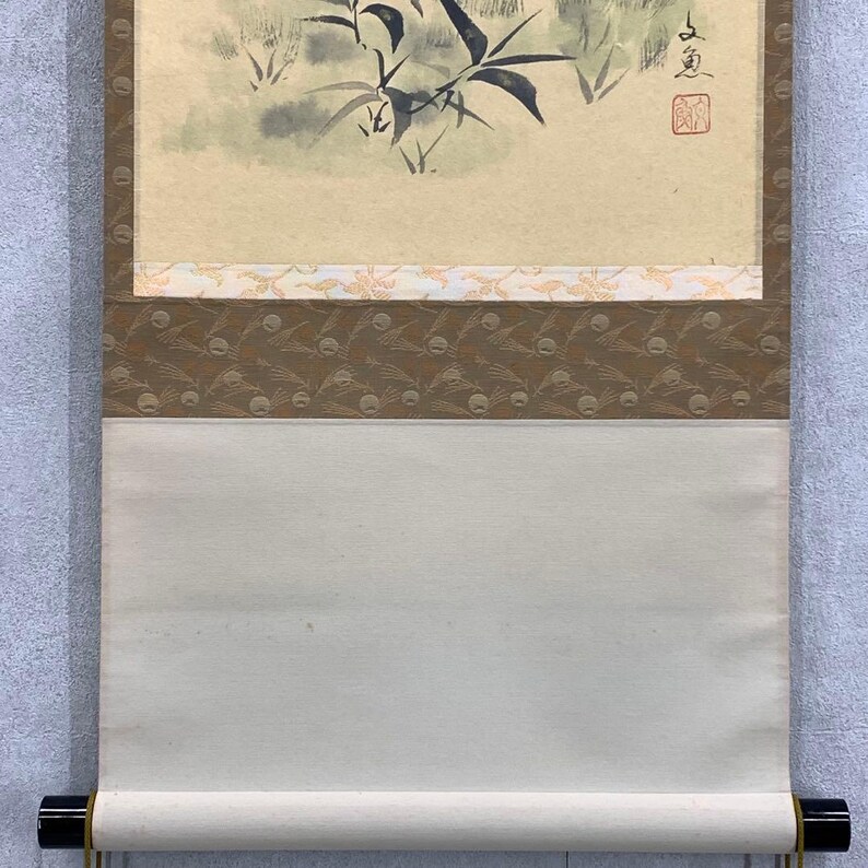 May include: Japanese scroll painting featuring a bamboo and foliage scene in muted green and gray tones. The artwork is mounted on a cream-colored paper with decorative borders in brown and gold. The scroll is finished with black wooden dowels.
