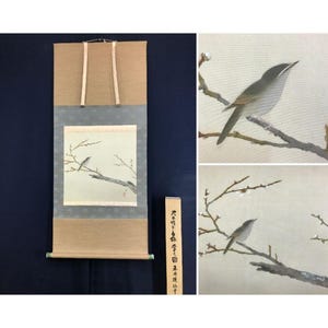 May include: A Japanese hanging scroll featuring a painting of a small grey bird perched on a bare branch with white blossoms. The scroll has tan fabric borders and a wooden storage box with black Japanese calligraphy text on the front panel.