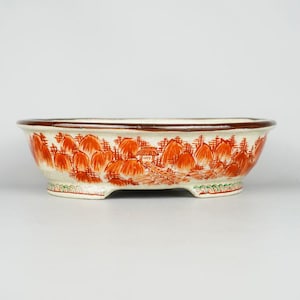 May include: Oval ceramic bonsai pot with a cream base and a dark brown rim. The pot features a hand-painted landscape design in orange and brown, with a small house and trees. The pot has three small feet.
