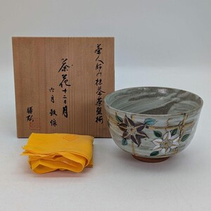 May include: A ceramic tea bowl with floral designs in purple, white, and gold, alongside a wooden box and a yellow cloth. The bowl features a gray and white marbled interior and a brown base. The box has Japanese text.