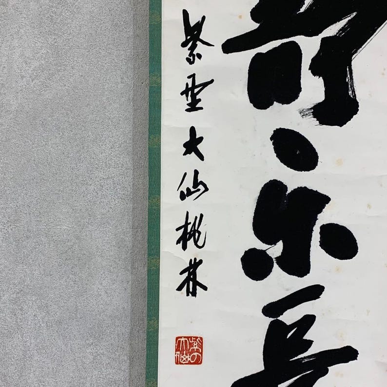 Puede incluir: Un pergamino vertical con caligraf&iacute;a japonesa en tinta negra sobre papel blanco. La caligraf&iacute;a incluye texto vertical a la izquierda y caracteres grandes y en negrita a la derecha. Un sello rojo es visible en la parte inferior. El pergamino tiene un borde verde a la izquierda.