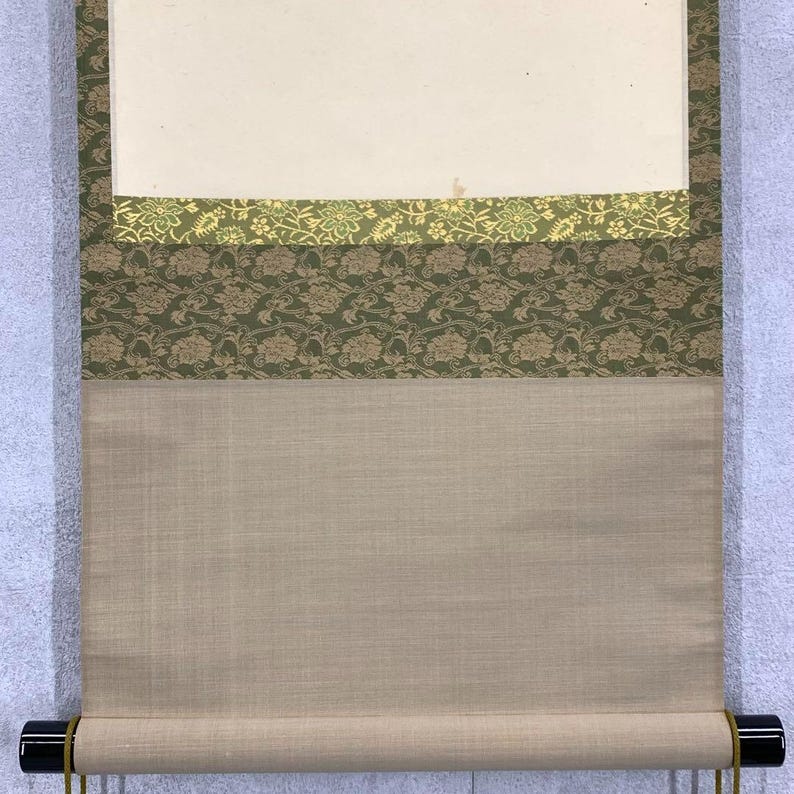 May include: A traditional Japanese scroll painting with a cream-colored paper section at the top. Below, there are layers of green and gold floral patterned fabric. The bottom section is a light beige fabric, and the scroll is held by a black rod.