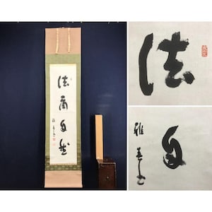 Op de afbeelding: Een verticale Japanse hangende rol met zwarte inkt kalligrafie op een witte papieren achtergrond. De rol heeft een lichtroze bovenrand en een groen patroon aan de onderkant. Twee close-up panelen tonen de zwarte inkttekens en rode stempels.