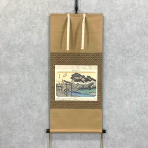 May include: A Japanese scroll painting depicting a landscape with a torii gate, mountains, and water. The artwork is framed by a brown patterned border and a tan background. The scroll is suspended by white cords.