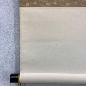 May include: A partially rolled scroll with a cream-colored paper surface. The top edge features a decorative band with a repeating pattern of gold and brown circular designs. The scroll is mounted on a black roller.