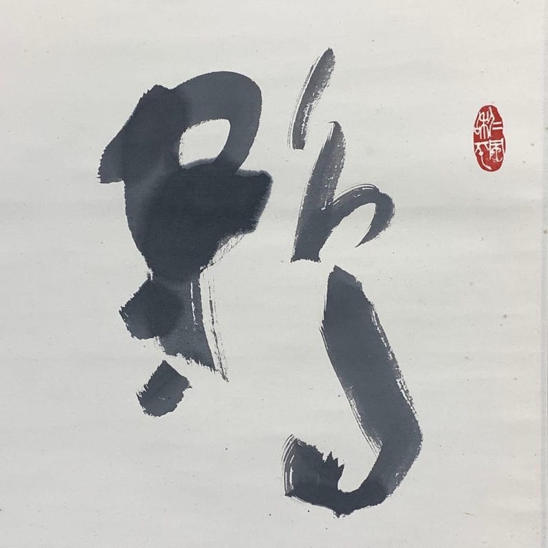 May include: Ink wash calligraphy on white paper. The dark gray brushstrokes form a stylized character, with a red seal in the upper right corner. The artwork has a minimalist aesthetic.
