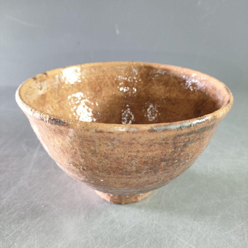 May include: A handmade, rustic ceramic bowl with a warm, earthy color palette. The bowl features a textured surface and a slightly flared rim. The exterior has a mottled brown appearance, while the interior has a glossy finish.