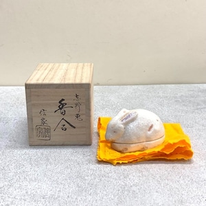 以下が含まれることがあります： 黄色い布の上に置かれた、蓋付きの陶器製のウサギの置物。隣には木箱があります。ウサギは白でピンクのアクセントがあります。木箱には日本語の文字が書かれています。