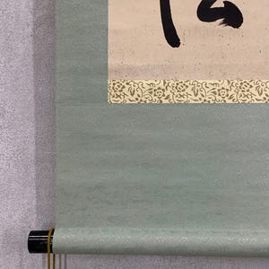 May include: A traditional Japanese scroll painting with black calligraphy on a cream-colored background. The scroll is bordered by a decorative floral pattern and a light green textured fabric. The bottom has a black roller with a gold cord.