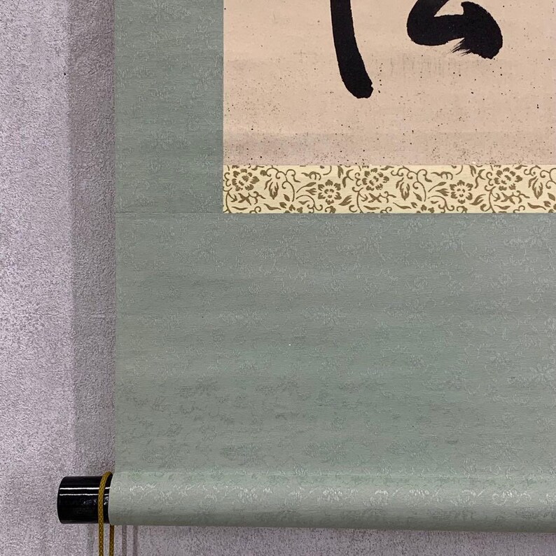 May include: A traditional Japanese scroll painting with black calligraphy on a cream-colored background. The scroll is bordered by a decorative floral pattern and a light green textured fabric. The bottom has a black roller with a gold cord.