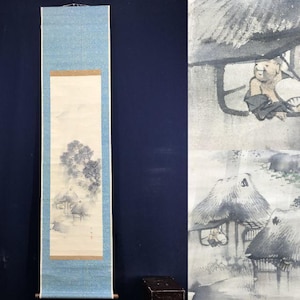Op de afbeelding: Een vintage Japanse rolschildering met een landschap met een boom, huizen en figuren. Het kunstwerk is voornamelijk in grijstinten en blauw, met een lichtblauwe rand en houten deuvels aan de onderkant.