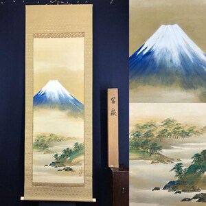 Pode incluir: Uma pintura japonesa em rolo retratando o Monte Fuji, com um pico coberto de neve e tons de azul e branco. A paisagem inclui árvores e água, em um fundo bege. O rolo tem uma borda decorativa e pinos de madeira.