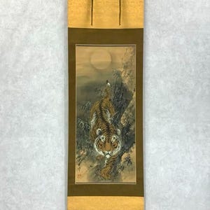 May include: A traditional Japanese scroll painting featuring a tiger descending a rocky hillside under a full moon. The artwork uses shades of brown, gold, and green, with the tiger's stripes and facial features clearly detailed.