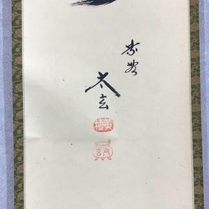 May include: A vertical scroll with Japanese calligraphy in black ink and red stamps. The calligraphy is on a cream-colored background, with a green and gold patterned border on the sides. The top of the scroll has a black ink brushstroke.